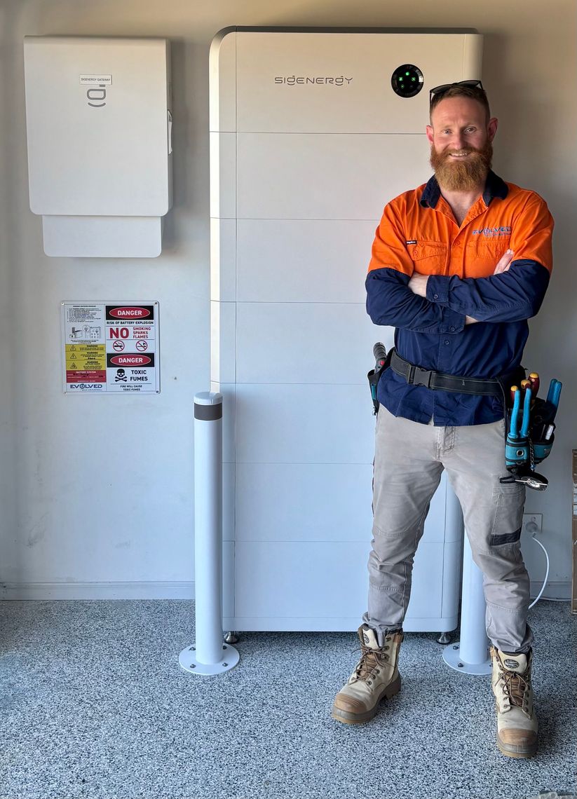 A man with a beard, wearing safety boots, tan work pants, and a long-sleeved orange and navy work shirt, standing with arms crossed in front of large industrial equipment. The equipment includes a white box with warnings and labels, and a sleek, silver rectangular device with green indicator lights.